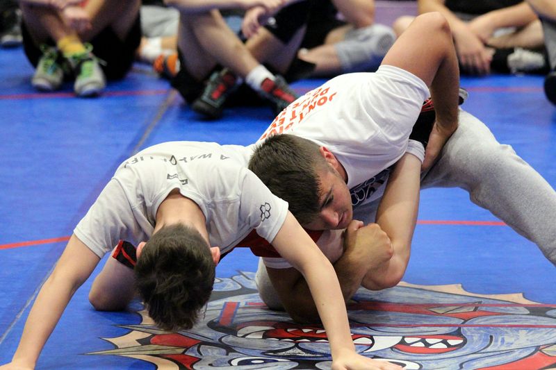 Two wrestlers in white shirts on a blue mat, one finishing a single leg at Ken Chertow's wrestling camp