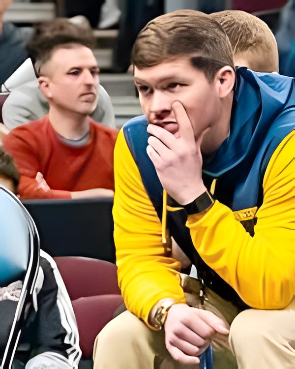 Mitchell Port seated matside in a gold and navy jacket, hand at his chin, Ken Chertow wrestling camp coach