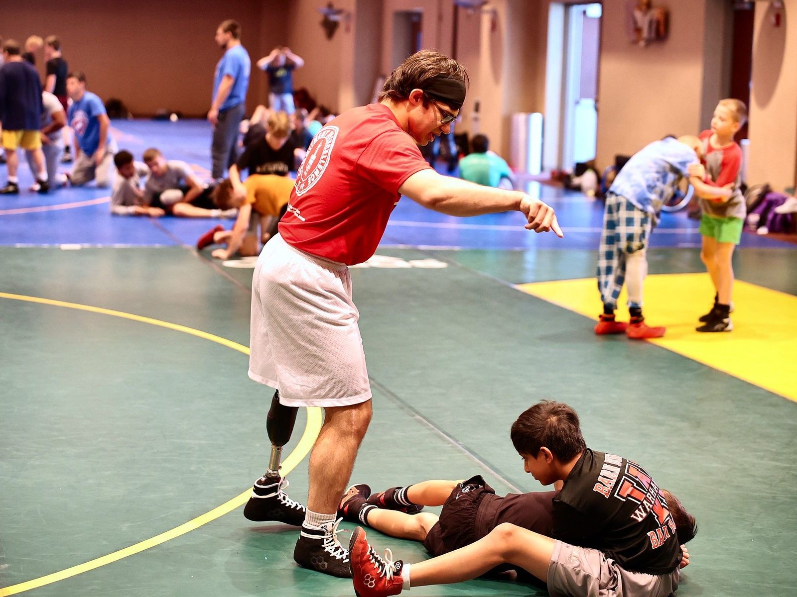 A coach in a red shirt showing young campers a move on the mat at youth wrestling camp