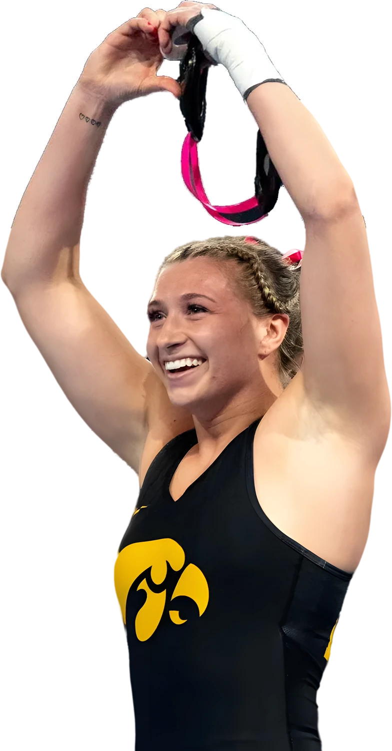 Kylie Welker in an Iowa singlet making a heart with her hands in front of the gold SuperGirl girls wrestling camp shield