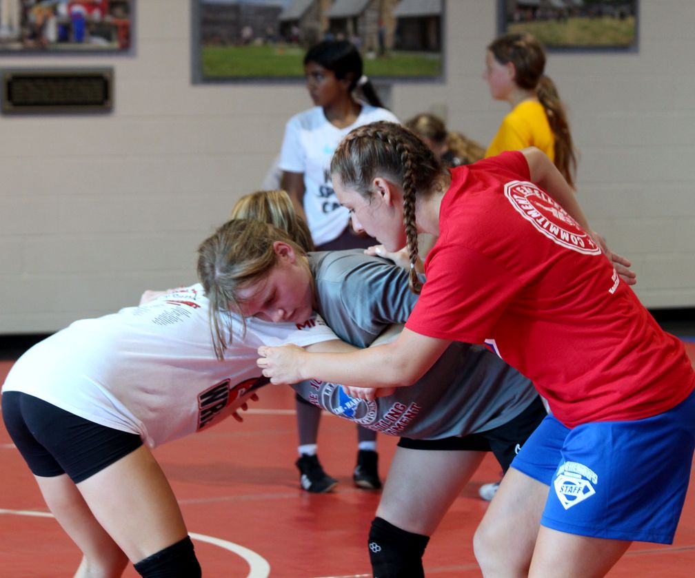 Girls hand fighting in a drill on a red mat at girls wrestling camp while other campers look on
